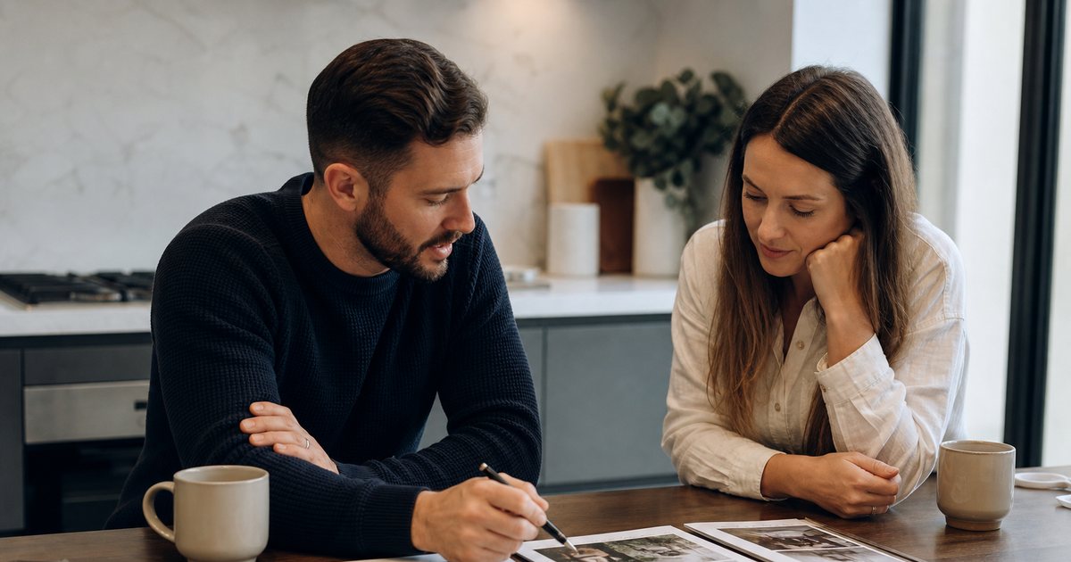 Engaged couple sitting at a kitchen table with three printed proposal folders spread out, coffee mugs, natural window light, one person holding a pen